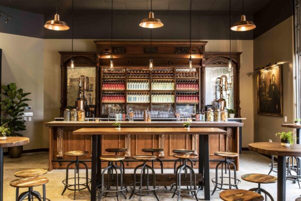 Elegant bar with a dark wooden counter, shelves of colorful bottles, distillation equipment, and industrial high-top seating.