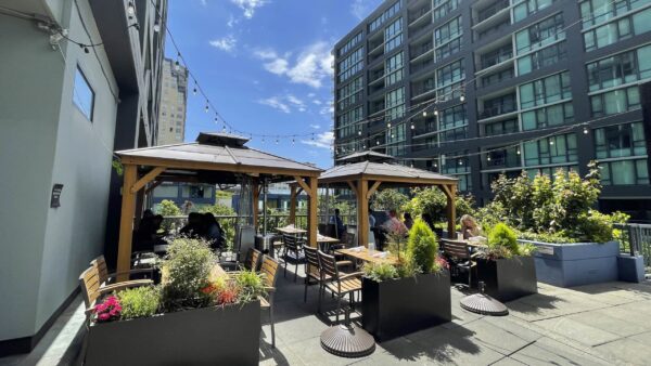 Outdoor restaurant patio with two wooden gazebos, tables, and diners. Overhead string lights, vibrant greenery, and a tall city building under a blue sky.