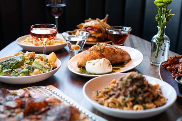 An appetizing spread of various dishes, including garlic bread with burrata, Caesar salad, pasta, wine, and cocktails.