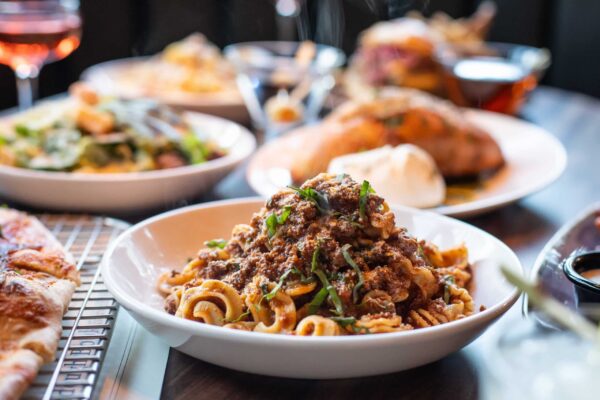 Steaming pasta with rich meat sauce and fresh basil in a bowl. A full table with plates of salad, pizza, and rosé wine.
