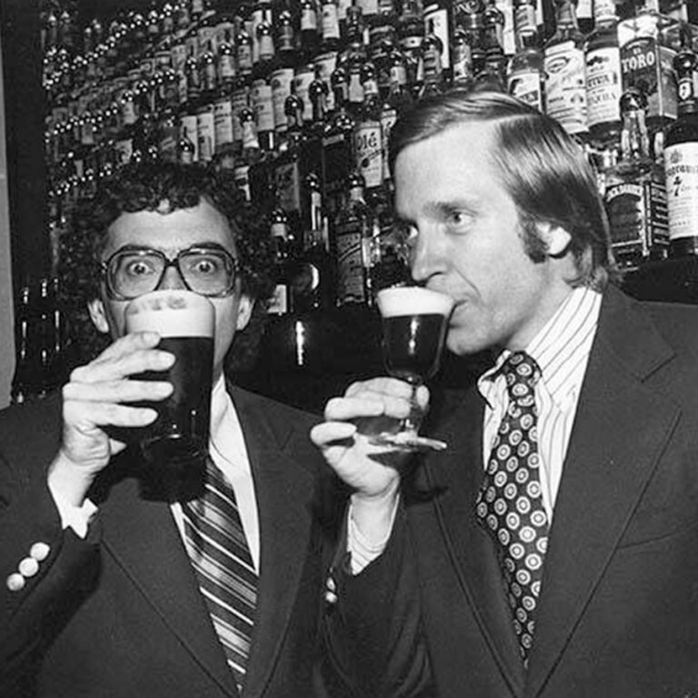 Tim and Mick in suits drinking beer with the wall of spirits behind them