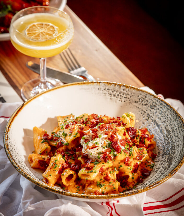 Bowl of rigatoni pasta in a spicy cream sauce topped with crispy bacon, burrata, and fresh chives, served alongside a yellow citrus cocktail in a coupe glass on a wooden table
