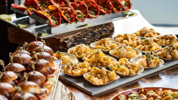 A vibrant buffet features red-shelled tacos on a stand, mini beef sliders with picks, and many small bowls of creamy pasta.