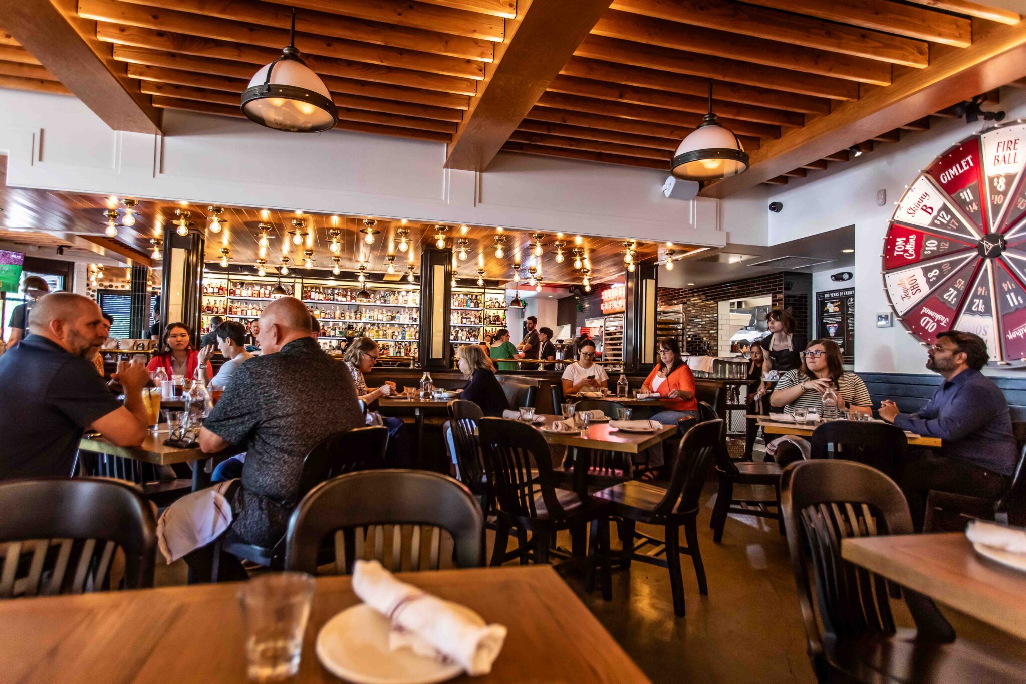 Busy dining room at Von's 1000Spirits Kirkland with guests seated at wooden tables, a fully stocked bar in the background, warm Edison bulb lighting, wood beam ceiling, and the Wagering Wheel visible on the right wall