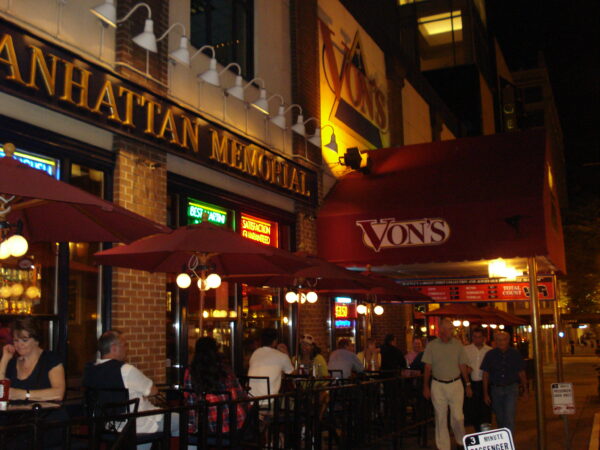 Exterior of the original Von’s at the Roosevelt Hotel in downtown Seattle, glowing at night with red awnings, neon signs, and guests dining on the sidewalk patio along 1st Avenue.