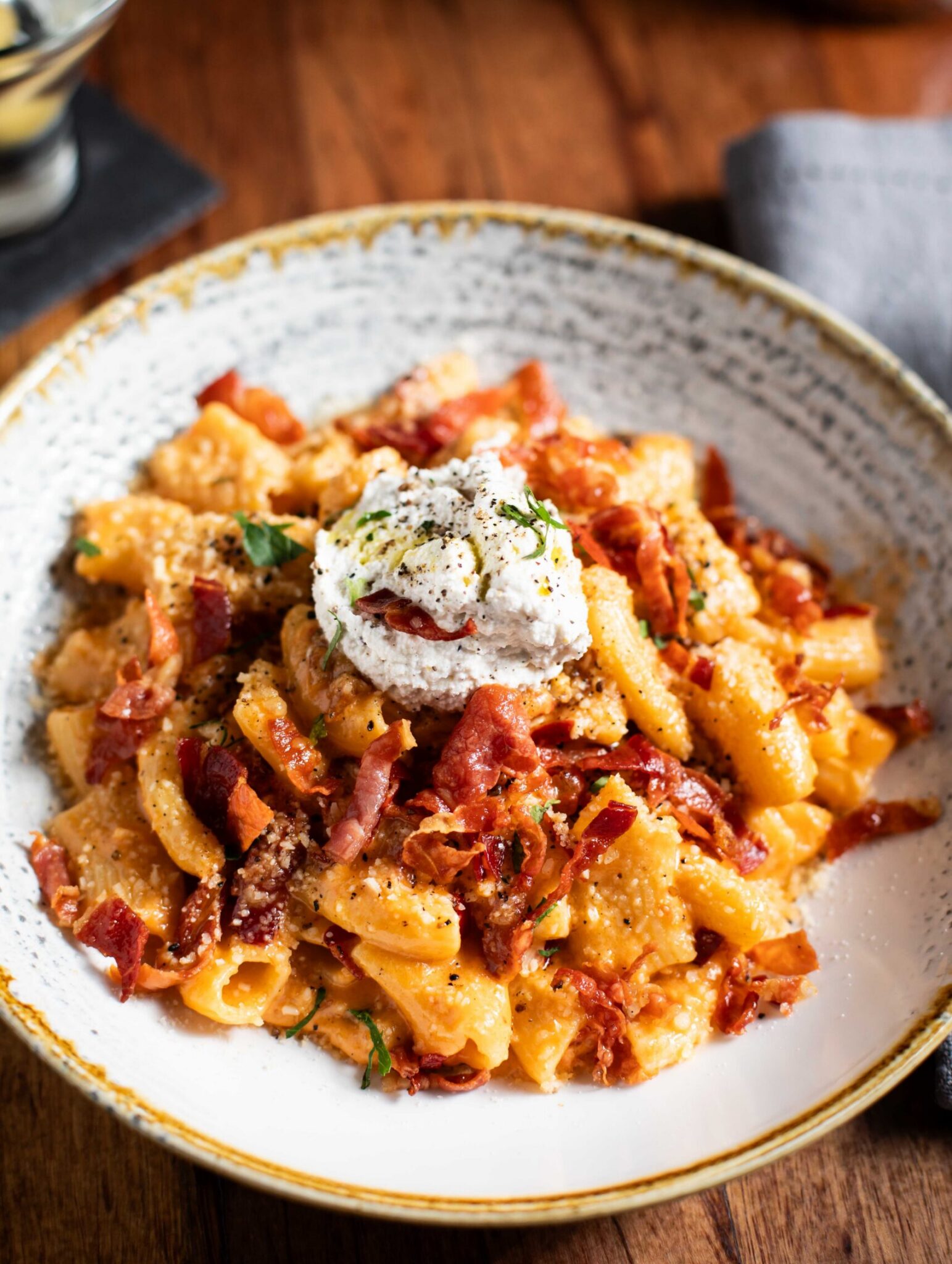 Rigatoni pasta topped with whipped ricotta, crispy bacon, and cracked black pepper in a rustic bowl.