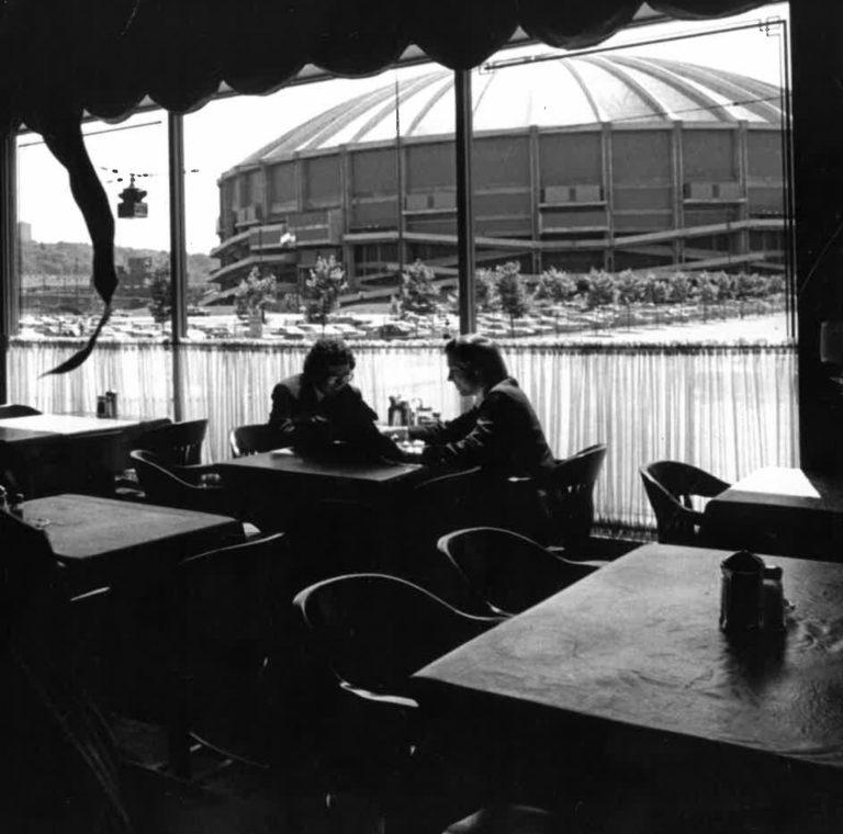 Black-and-white photo of two men seated at a table inside a restaurant, silhouetted against large windows with the Seattle Kingdome visible in the background.