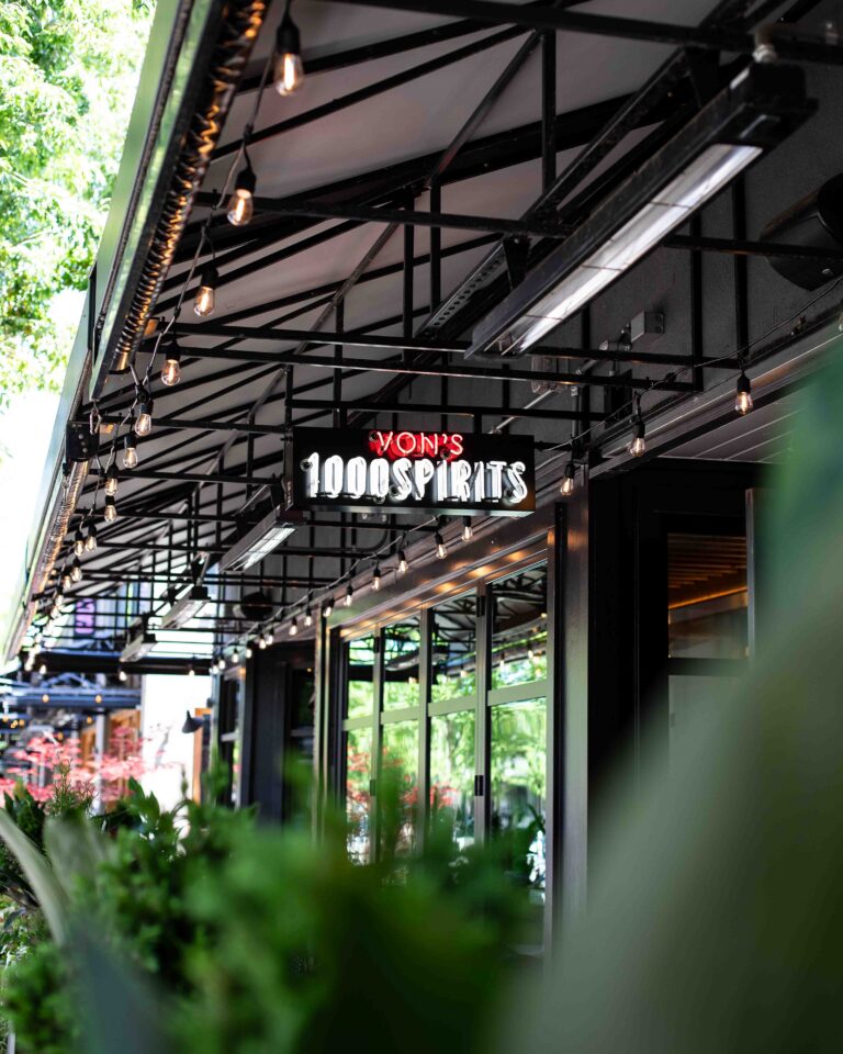 Exterior covered patio of Von's 1000Spirits Kirkland with a glowing neon sign, Edison string lights, black steel awning, and lush green landscaping in the foreground