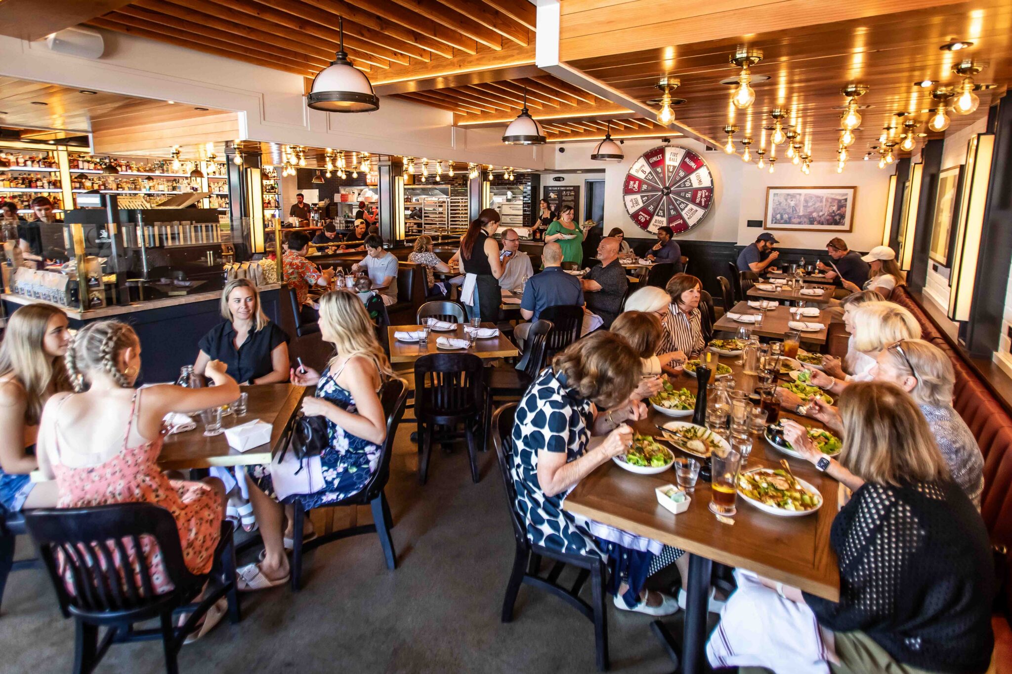 Busy dining room at Von's 1000Spirits Kirkland with guests seated at wooden tables, a server taking orders, the Wagering Wheel visible on the wall, fully stocked bar in the background, and warm Edison bulb lighting under a wood beam ceiling