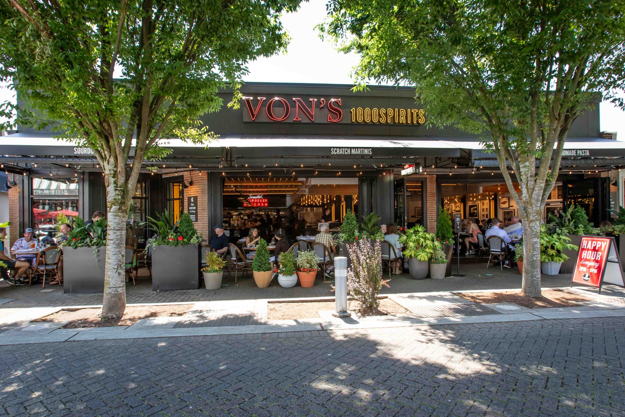 Outdoor patio at Von's 1000Spirits Kirkland with guests dining at bistro chairs among lush green planters and flowering pots, red neon signage visible above the entrance, a Happy Hour sidewalk sign to the right, and tree-lined brick walkway in the foreground